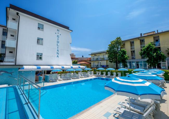 Piscina all'aperto con ombrelloni blu e hotel sullo sfondo.