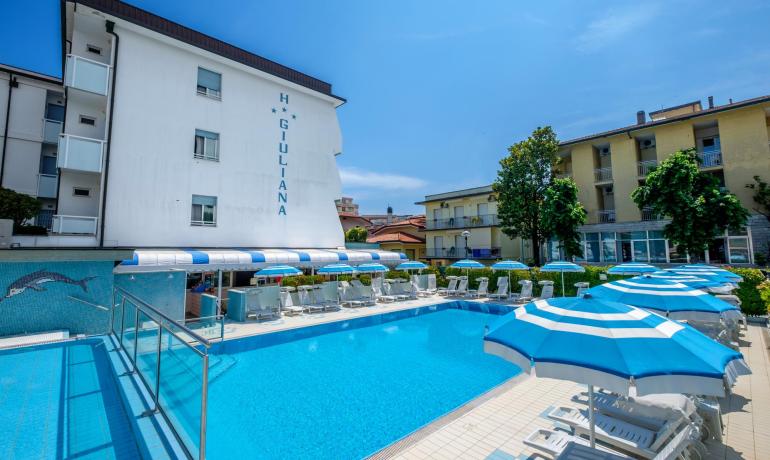 Piscina all'aperto con ombrelloni blu e hotel sullo sfondo.