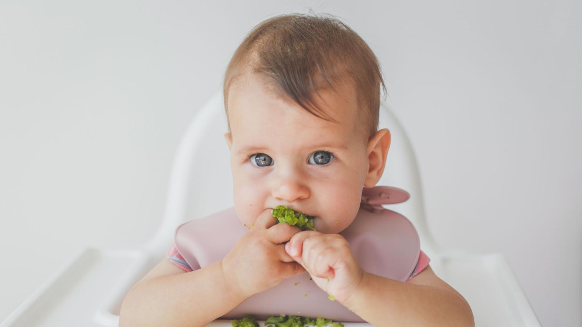 Bambino mangia broccoli con bavaglino rosa.