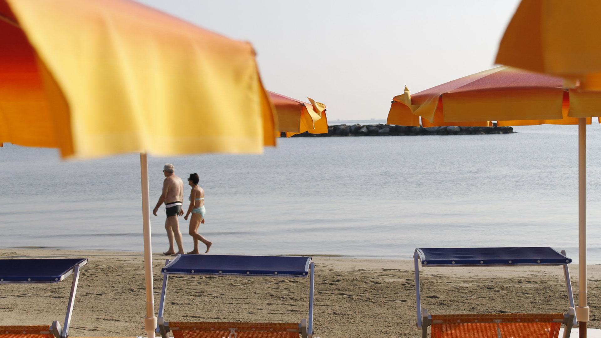 Spiaggia con ombrelloni arancioni, coppia cammina sul bagnasciuga.