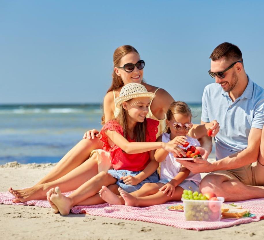 Famiglia felice fa picnic sulla spiaggia con frutta e sole.